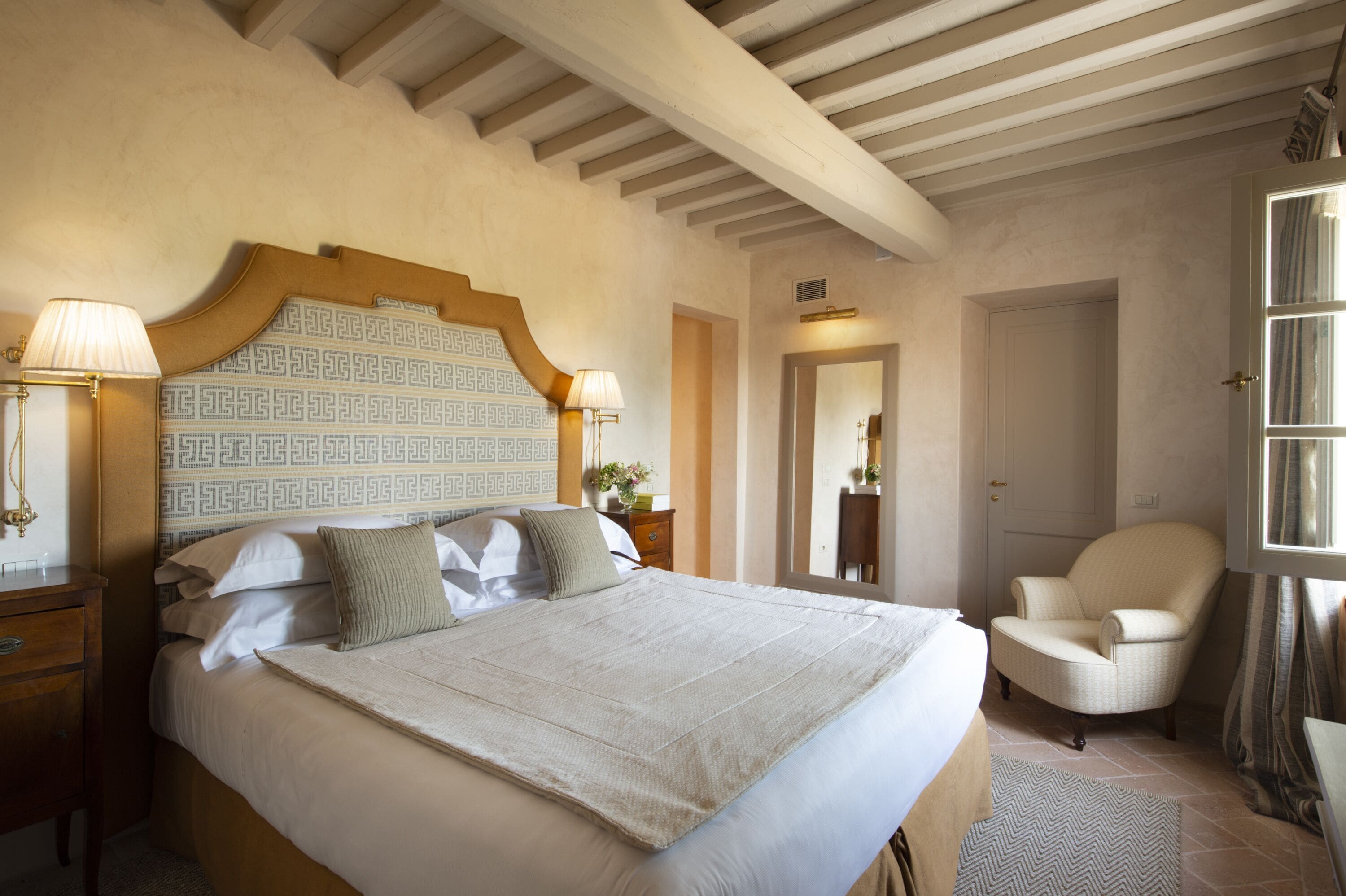 Une chambre à coucher avec un grand lit, une tête de lit en bois, deux tables de chevet avec des lampes, une chaise et une fenêtre avec des rideaux.