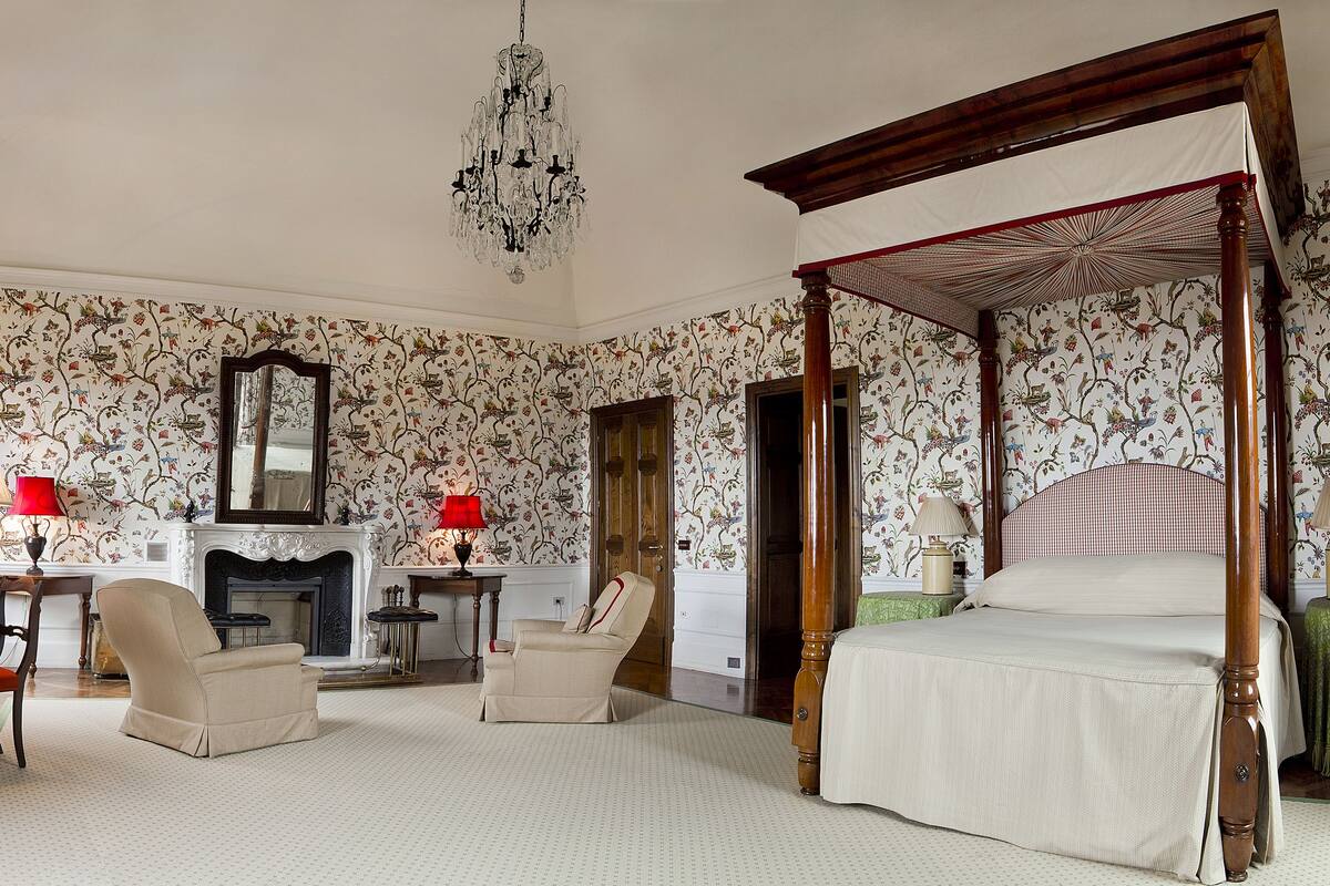 A bedroom with floral wallpaper, a four-poster bed, a fireplace, and a chandelier.