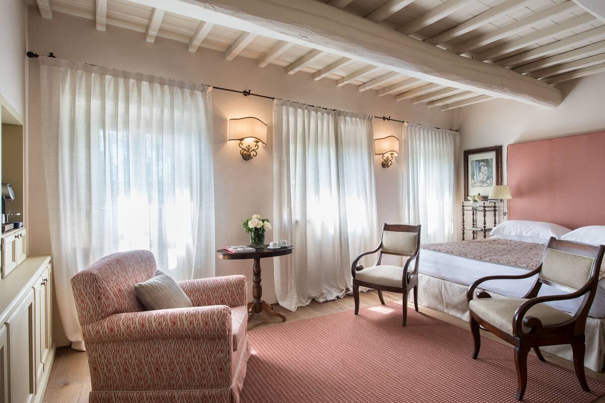 Habitación de hotel con cama, dos sillas, una mesita con un florero y un ventanal con cortinas.
