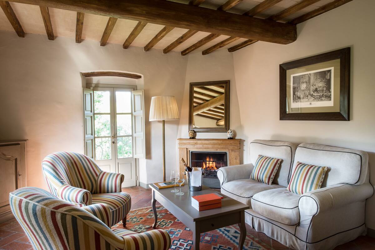 A cozy living room with a fireplace, striped armchairs, a sofa, and a coffee table with books and glasses.