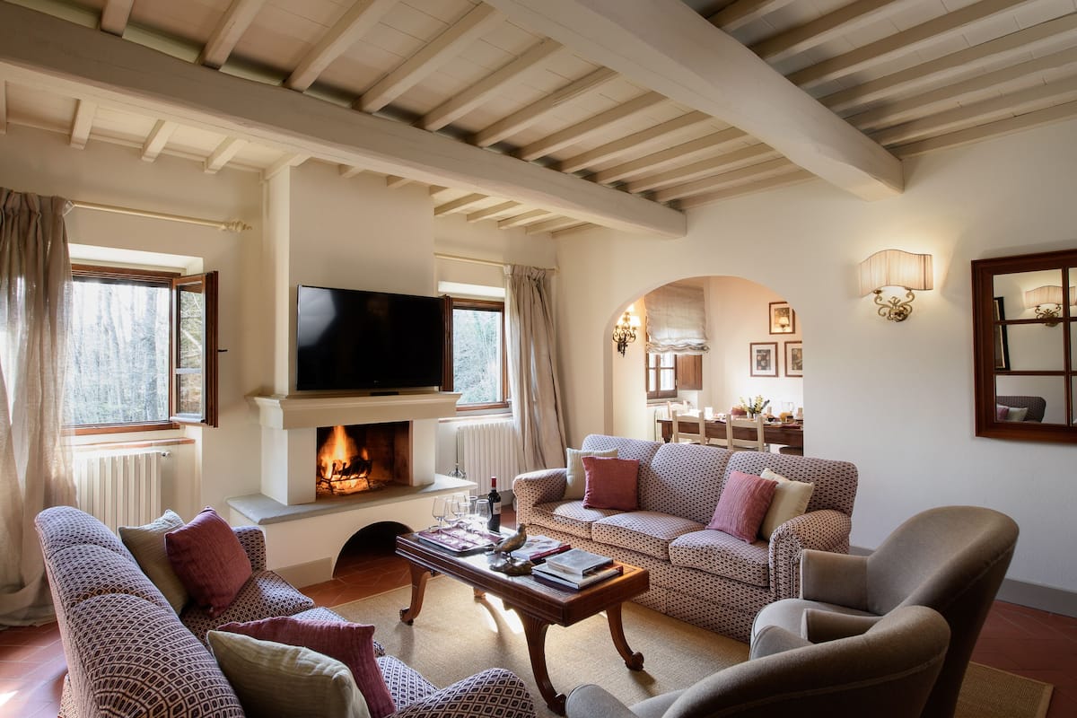 Un salón acogedor con sofás estampados, una chimenea, un televisor y un comedor con candelabro.