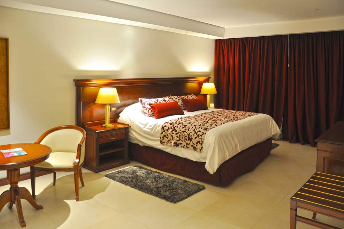 A hotel room with a bed, two bedside lamps, a chair, a small table, and a desk.