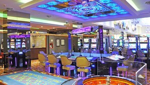 Un casino con múltiples máquinas tragamonedas, una mesa de ruleta y zonas de bar.