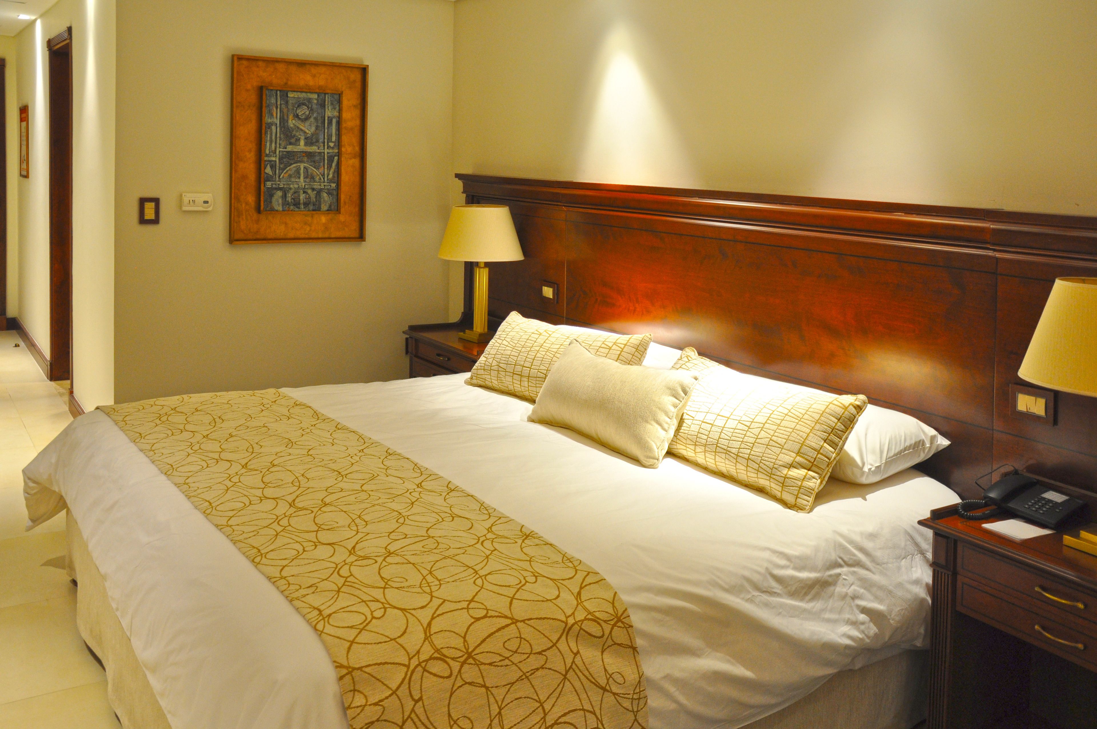 A hotel room with a large bed, a wooden headboard, a bedside table with a lamp, and a framed picture on the wall.