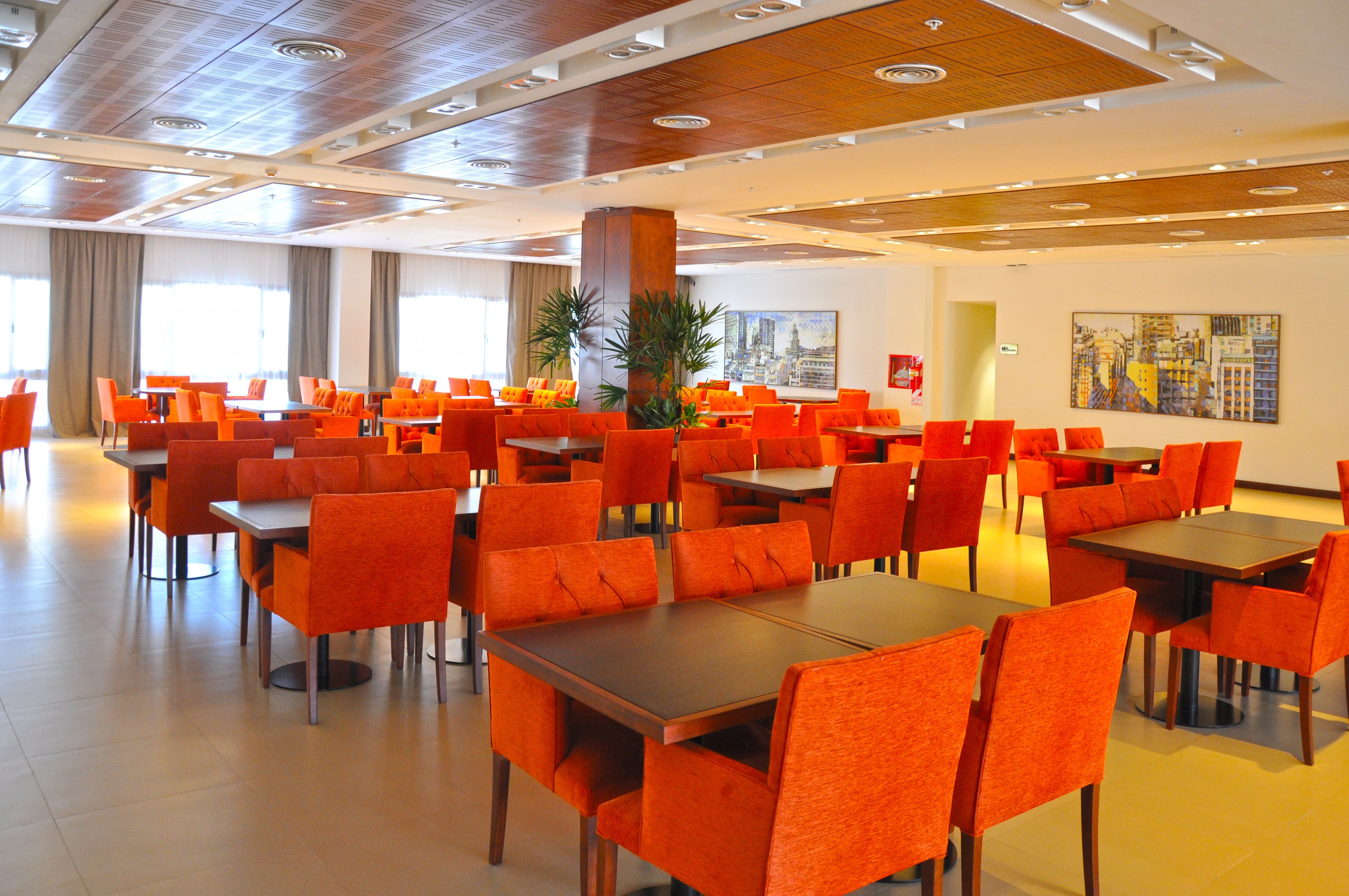 Een ruime eethoek met meerdere tafels en oranje stoelen, met een modern plafondontwerp en grote ramen.