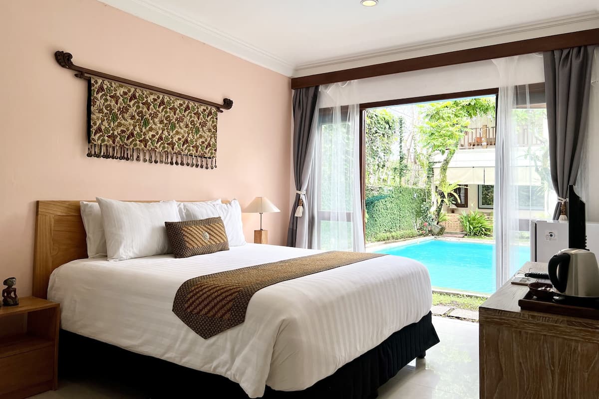 A bedroom with a large bed, a nightstand, a lamp, a decorative wall hanging, and a view of a swimming pool.
