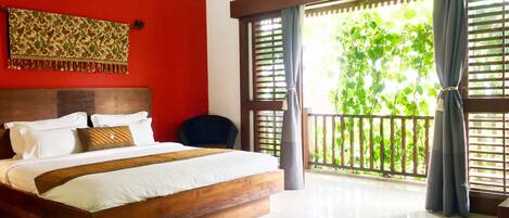 A hotel room with a large bed, a wooden headboard, a red accent wall, and a balcony with a view of greenery.