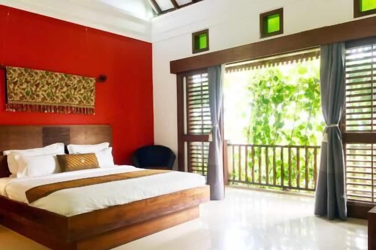 A bedroom with a wooden bed, a red accent wall, a view of greenery through large windows, and a ceiling with skylights.