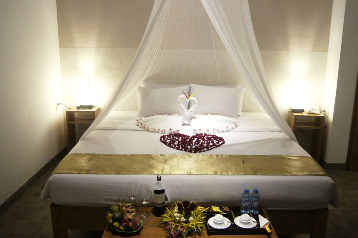 Una cama bien hecha con una decoración con forma de corazón, una mesita de noche con una botella y vasos, y una bandeja con botellas de agua y un plato.