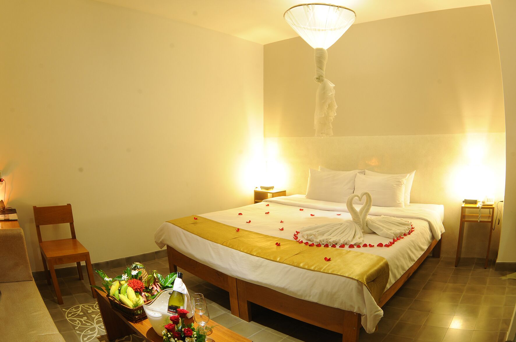 Una habitación de hotel con cama, mesitas de noche, una silla, una mesa con flores y una botella de vino.
