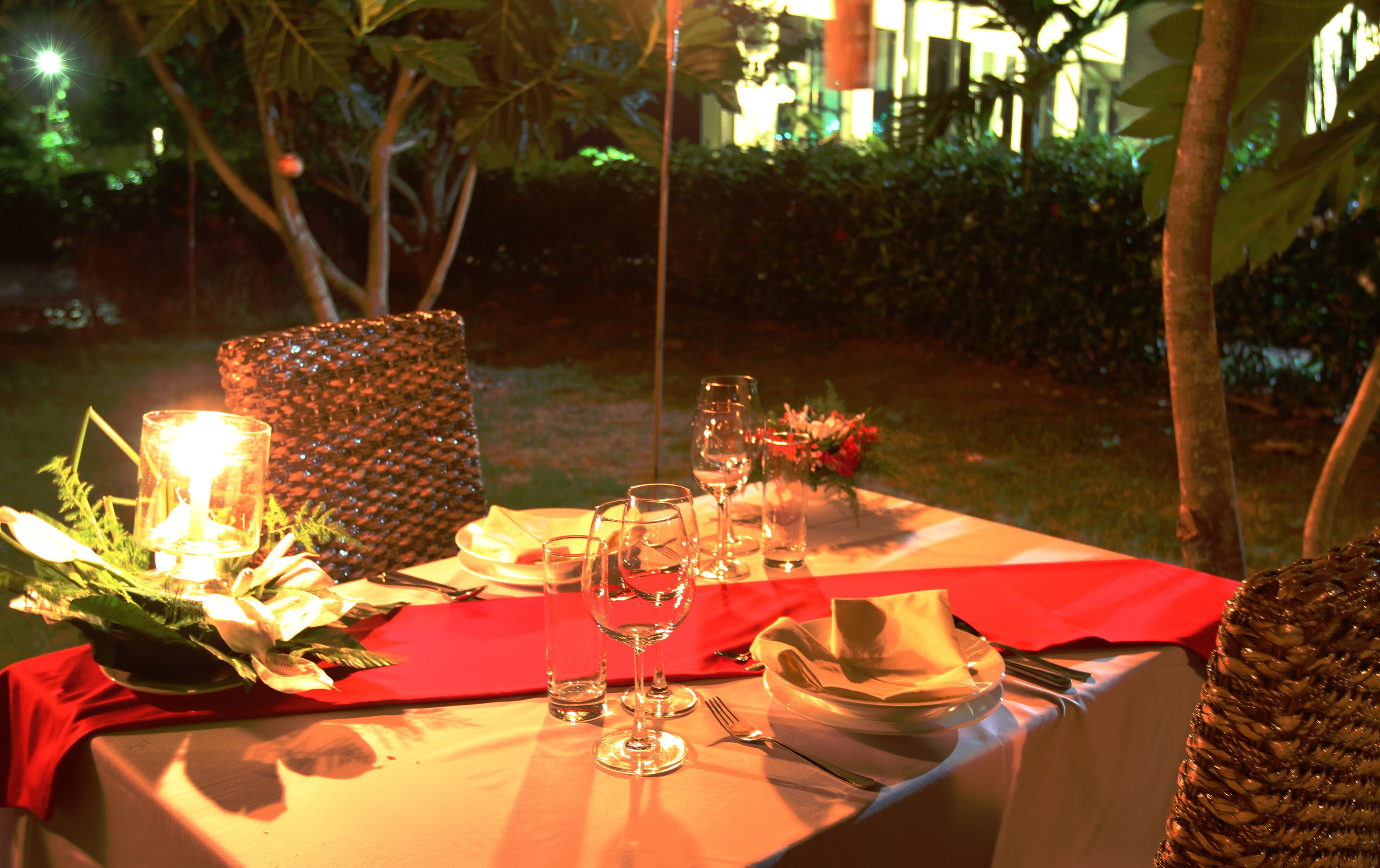 An outdoor dining setup with a table set for two, featuring a red tablecloth, glassware, and a candle centerpiece.
