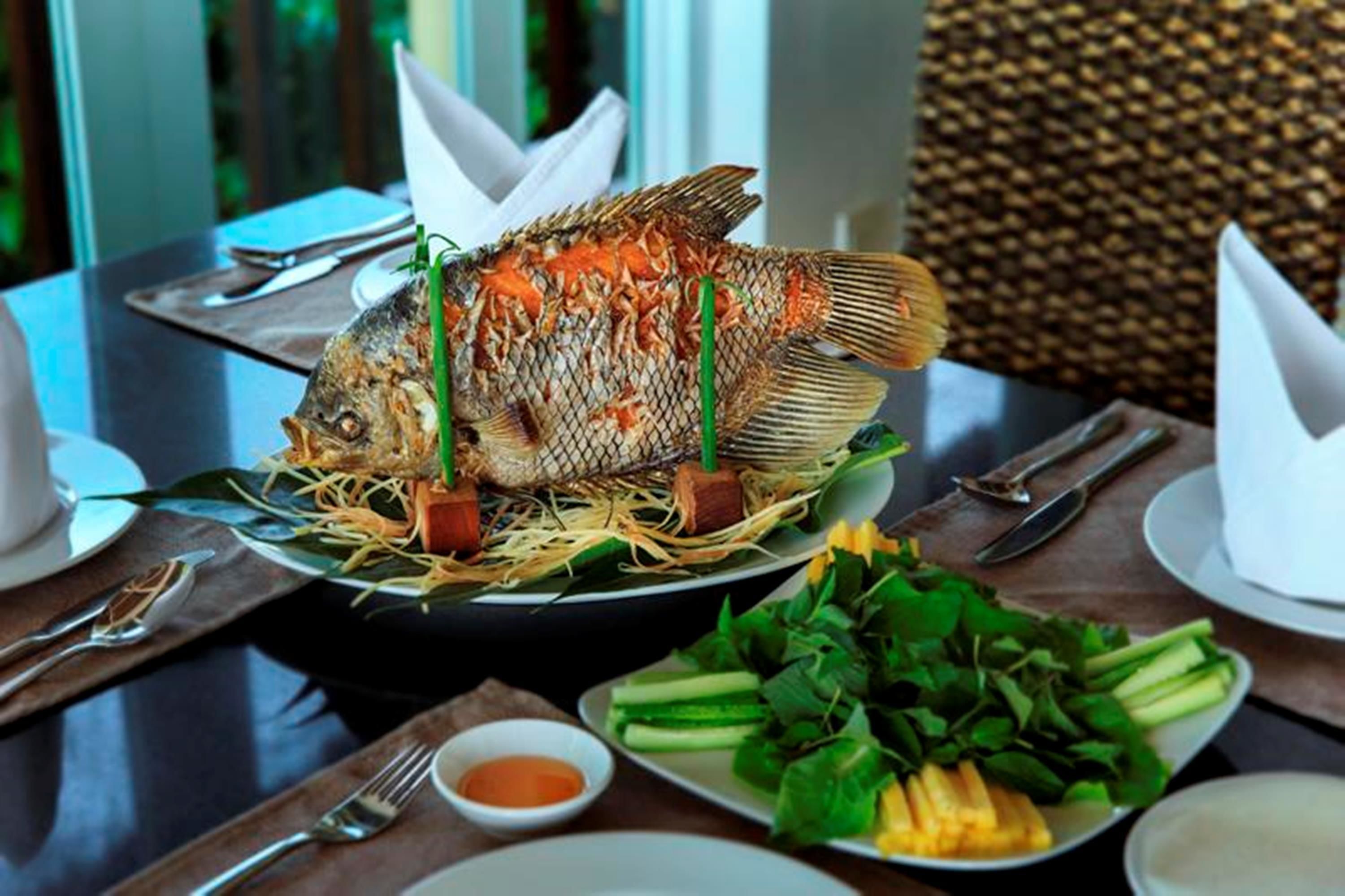 A dining table with a whole fish, vegetables, and a small bowl of sauce.
