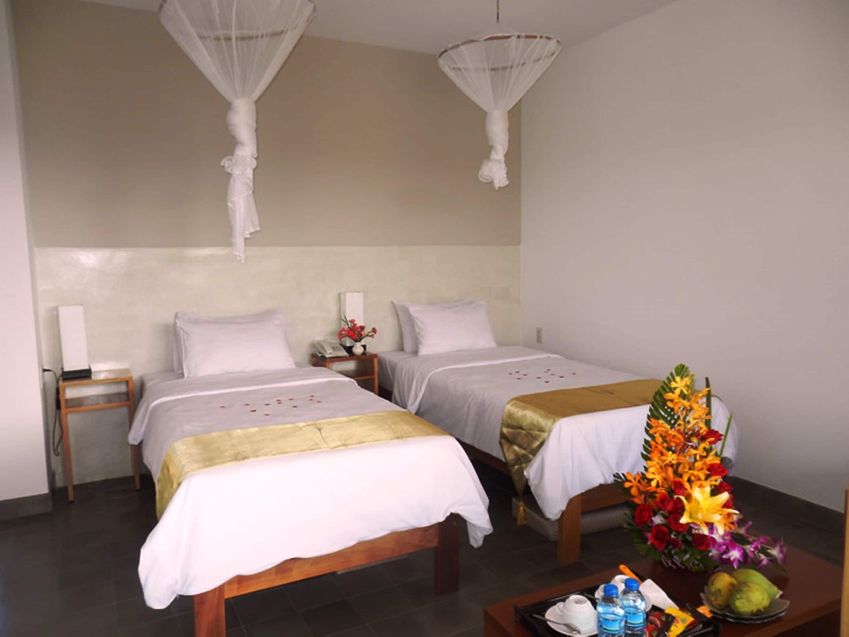A hotel room with two beds, a bedside table with a vase of flowers, and a nightstand with a lamp.