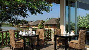 A dining area with wicker chairs and tables, overlooking a lake with thatched-roof buildings.