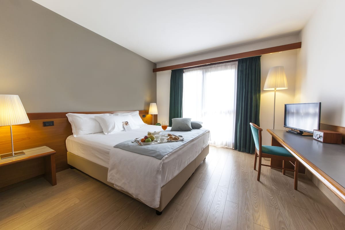 A hotel room with a bed, bedside table, lamp, TV, and a desk with a chair.