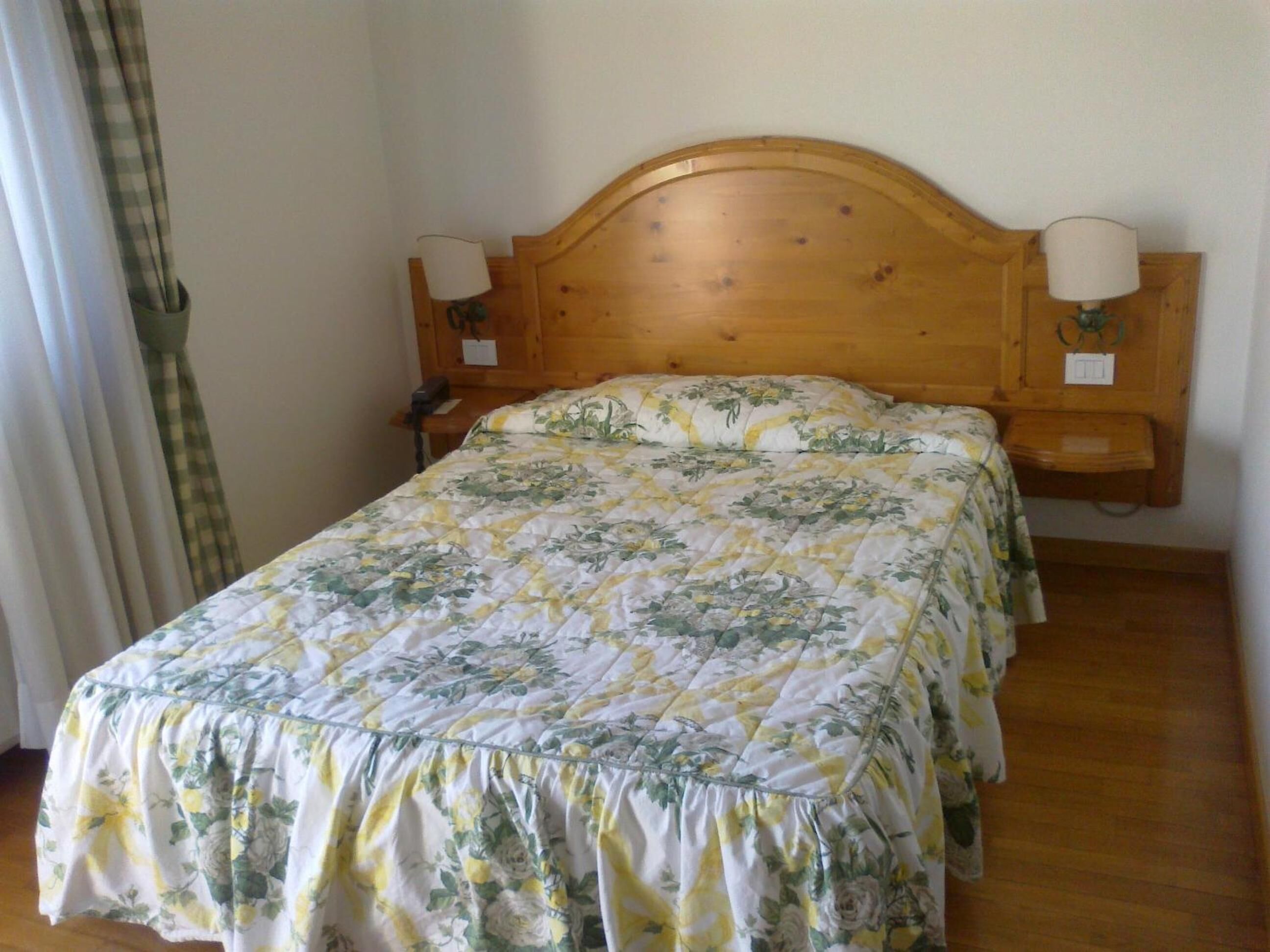 A single bed with a floral bedspread, wooden headboard, two wall-mounted lamps, and a small wooden nightstand.