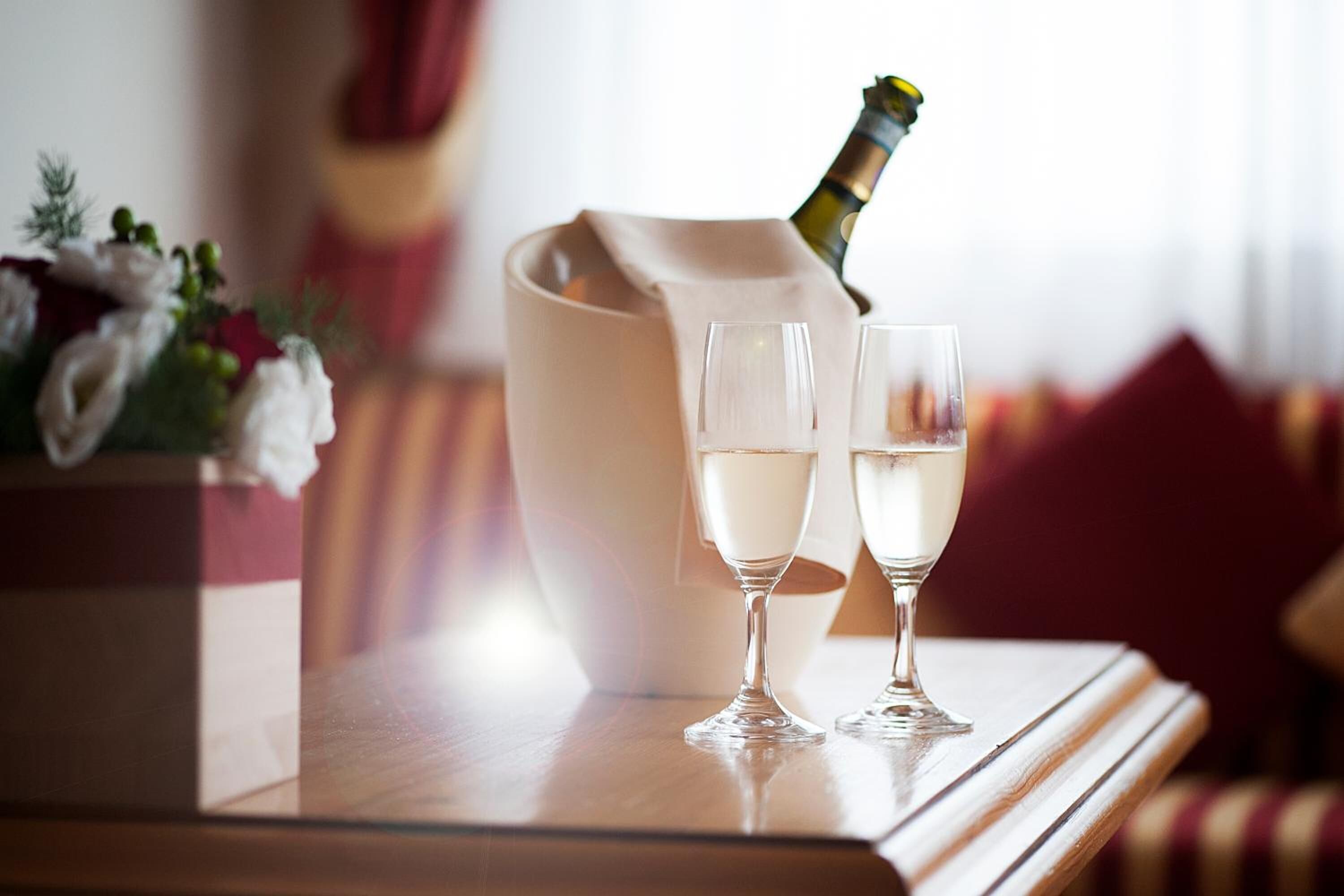 A champagne bottle being opened, two glasses, and a floral arrangement on a wooden table.