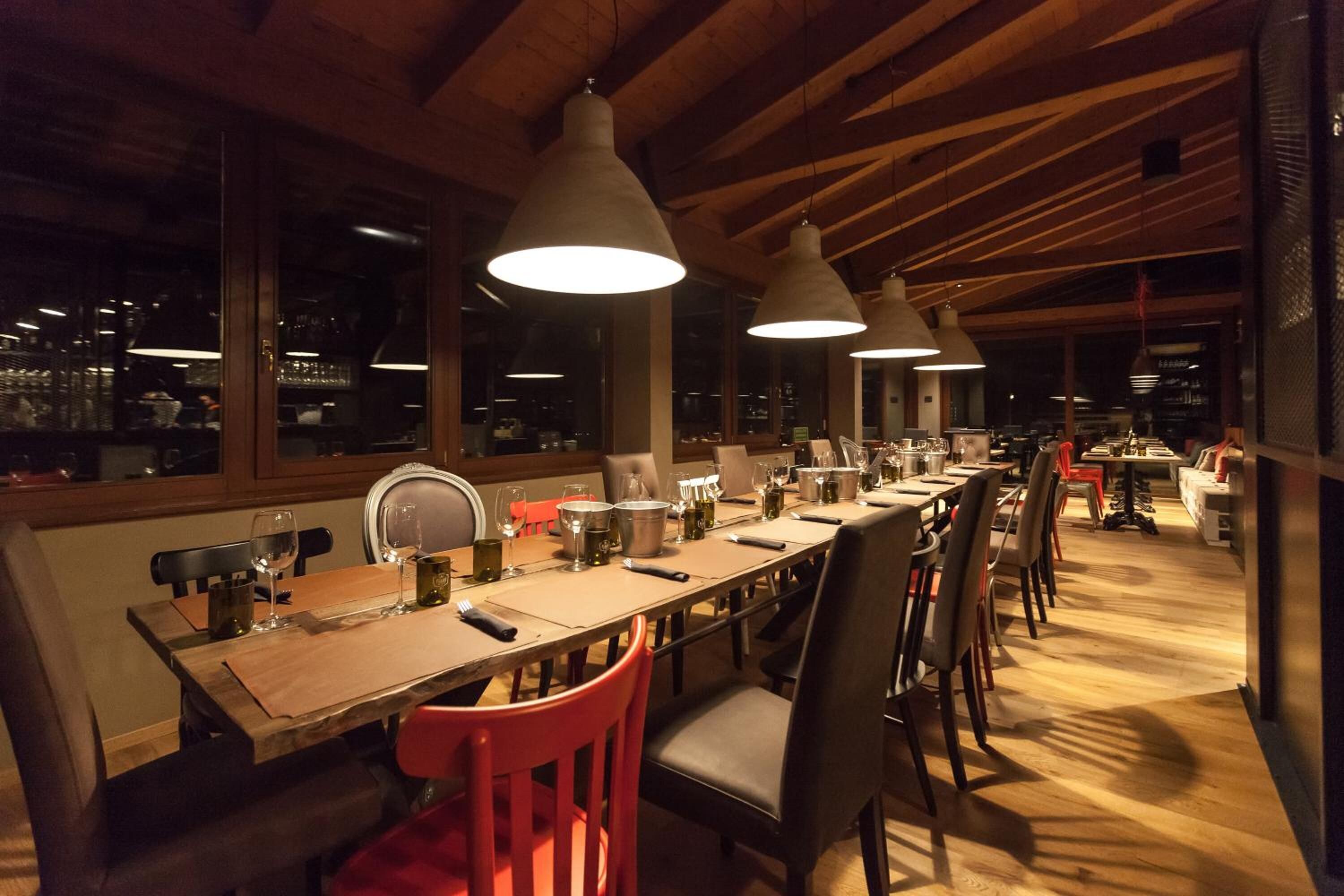 A long dining table with multiple chairs, overhead pendant lights, and a view of the kitchen area through large windows.