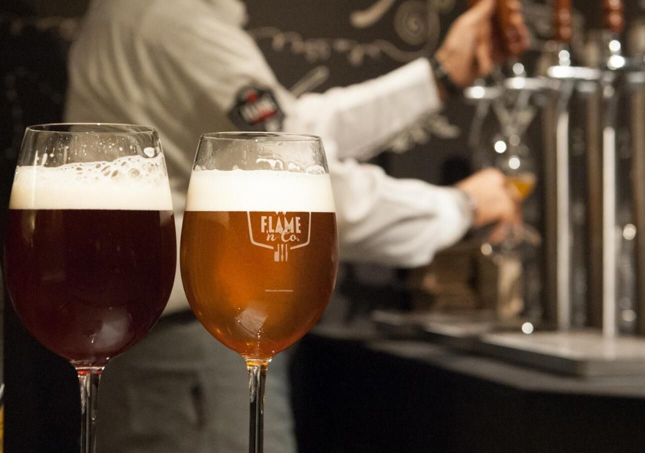 Dos vasos de cerveza con el logotipo "Flame n Co." sobre una barra.