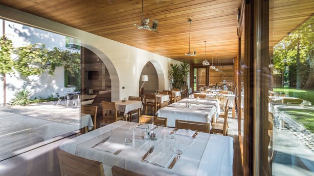 Un restaurant doté d’un plafond en bois et de fenêtres en arc de cercle, avec des tables dressées pour le service et une vue sur l’extérieur.