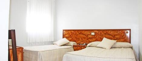 A hotel room with two beds, a wooden headboard, a bedside table, a chair, and a window with curtains.