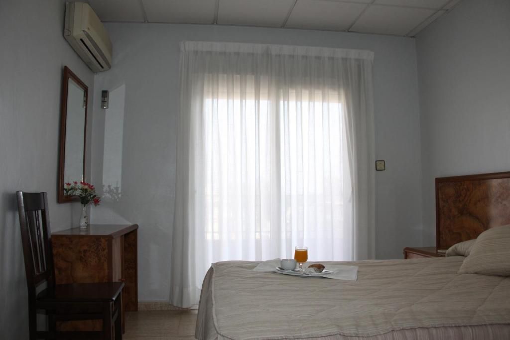 A bedroom with a bed, a bedside table, a chair, a mirror, and a window with curtains.