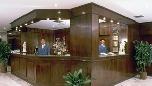 Un mostrador de recepción con dos hombres detrás, un reloj en la pared y plantas en macetas.