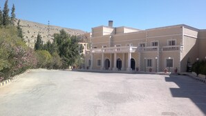 Un edificio grande con varios balcones y puertas arqueadas, rodeado de árboles y un paisaje montañoso.