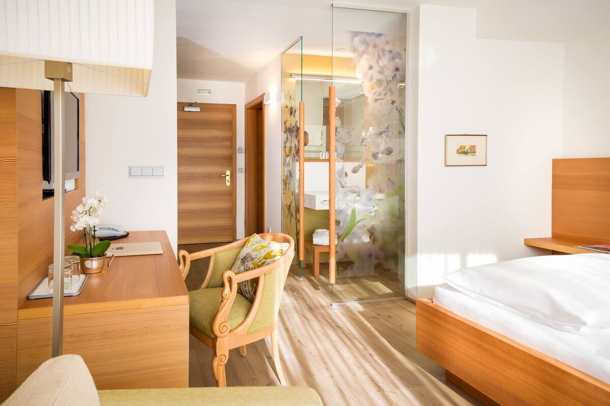 Une chambre d’hôtel avec un lit en bois, un bureau avec un téléphone et une plante, une chaise, un miroir et une salle de bain avec une porte en verre.