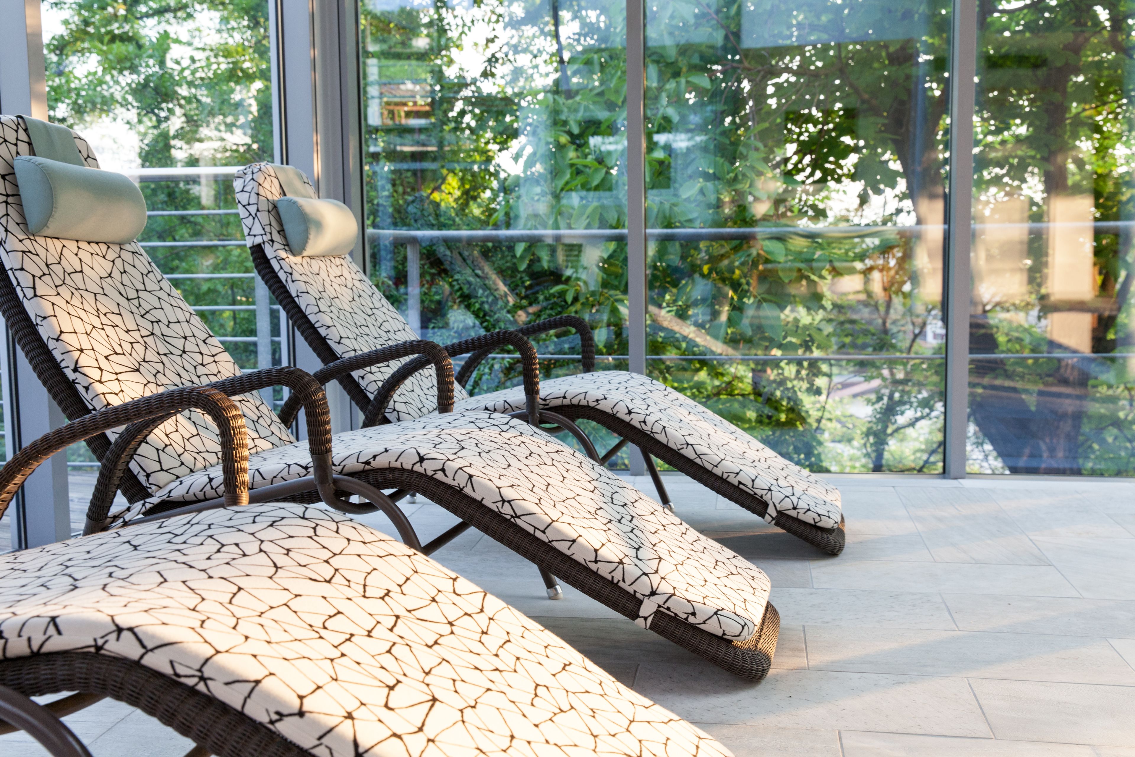 File di lettini prendisole con cuscini decorati, disposti in una veranda luminosa con ampie finestre che si affacciano su un paesaggio verde.