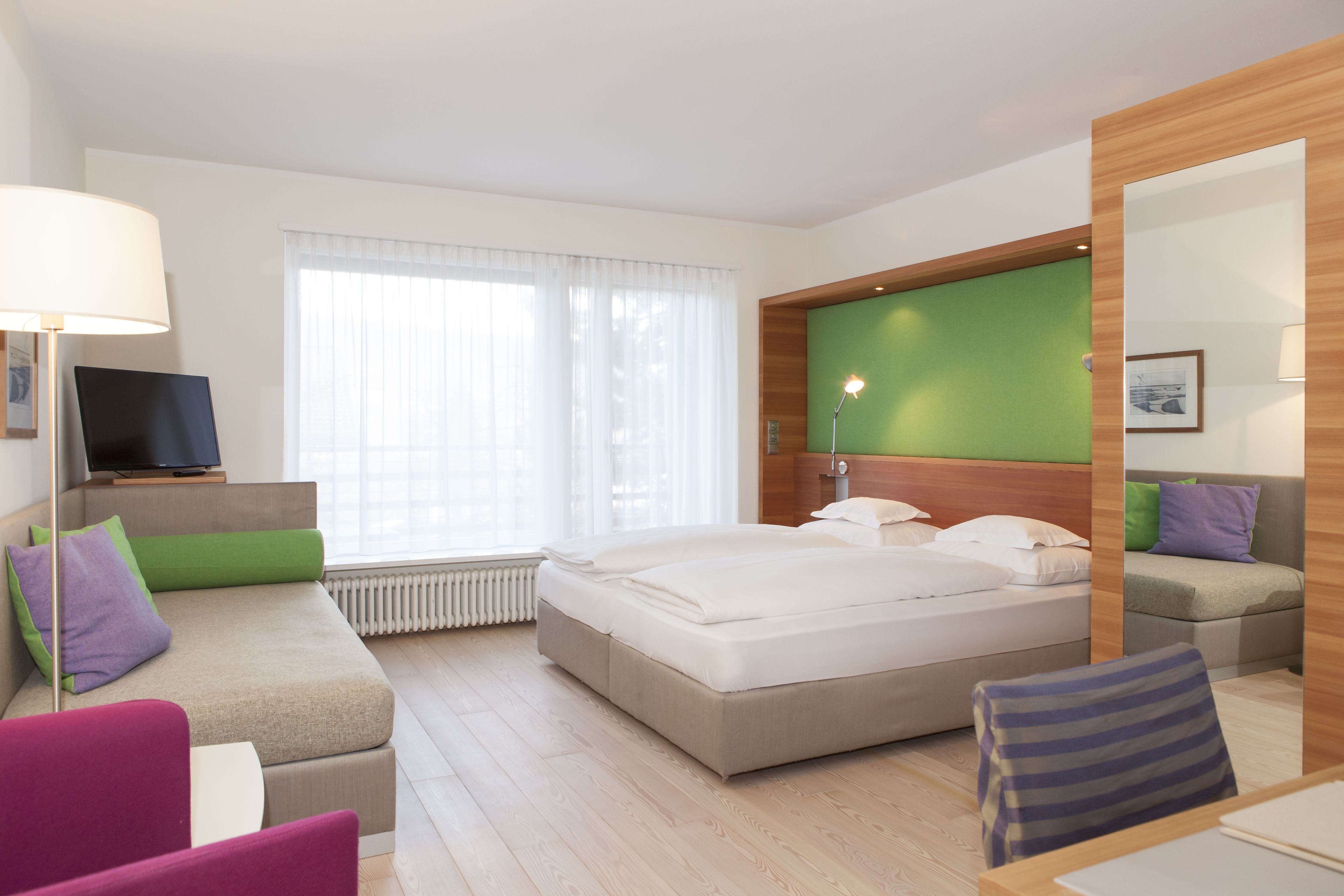Habitación de hotel con cama, sofá, sillón, televisor y ventana con cortinas.