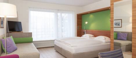 Habitación de hotel con cama, sofá, sillón, televisor y ventana con cortinas.