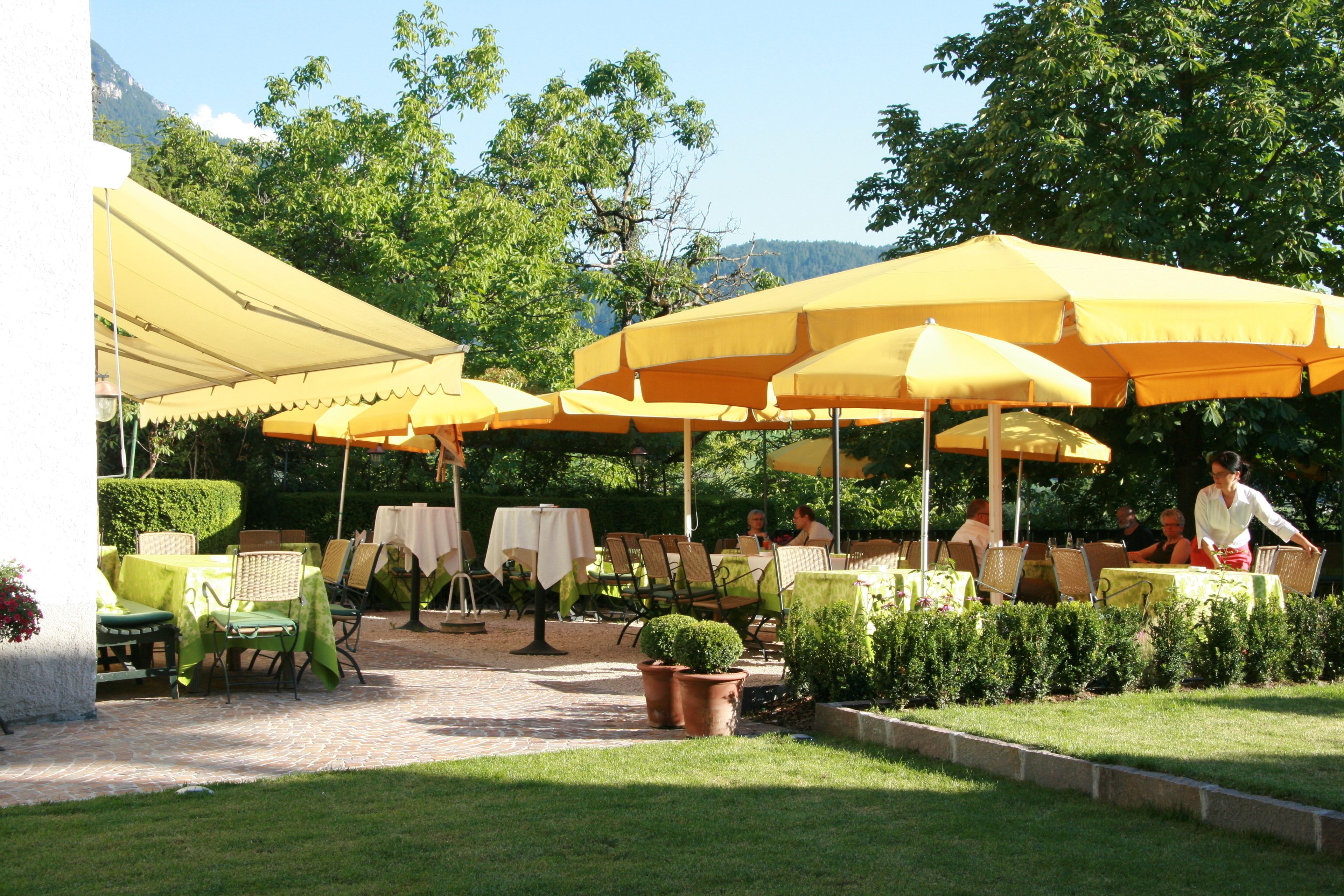 Een terras buiten met gele parasols, tafels en stoelen, omgeven door groen en een berg op de achtergrond.