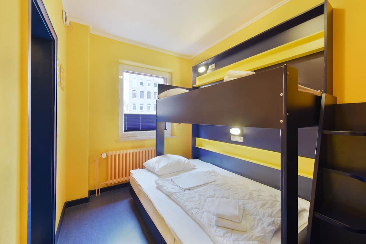 A compact hotel room with a bunk bed, a window with blinds, and a radiator.