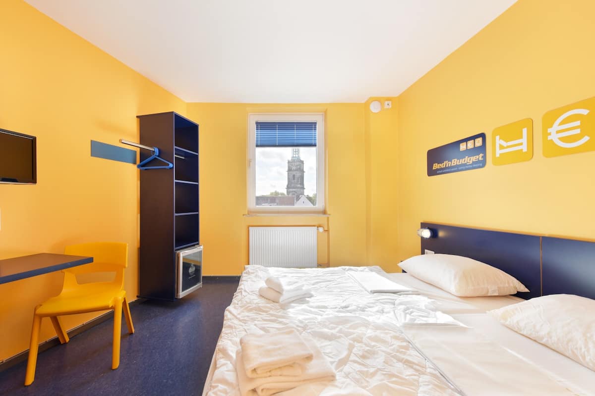 A hotel room with a bed, a desk, a chair, a wardrobe, and a window with a cityscape view.