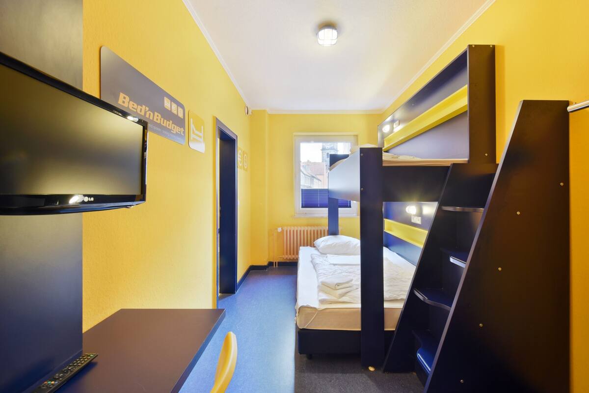 A hotel room with a bunk bed, a flat-screen TV mounted on the wall, a desk with a chair, and a window with blinds.