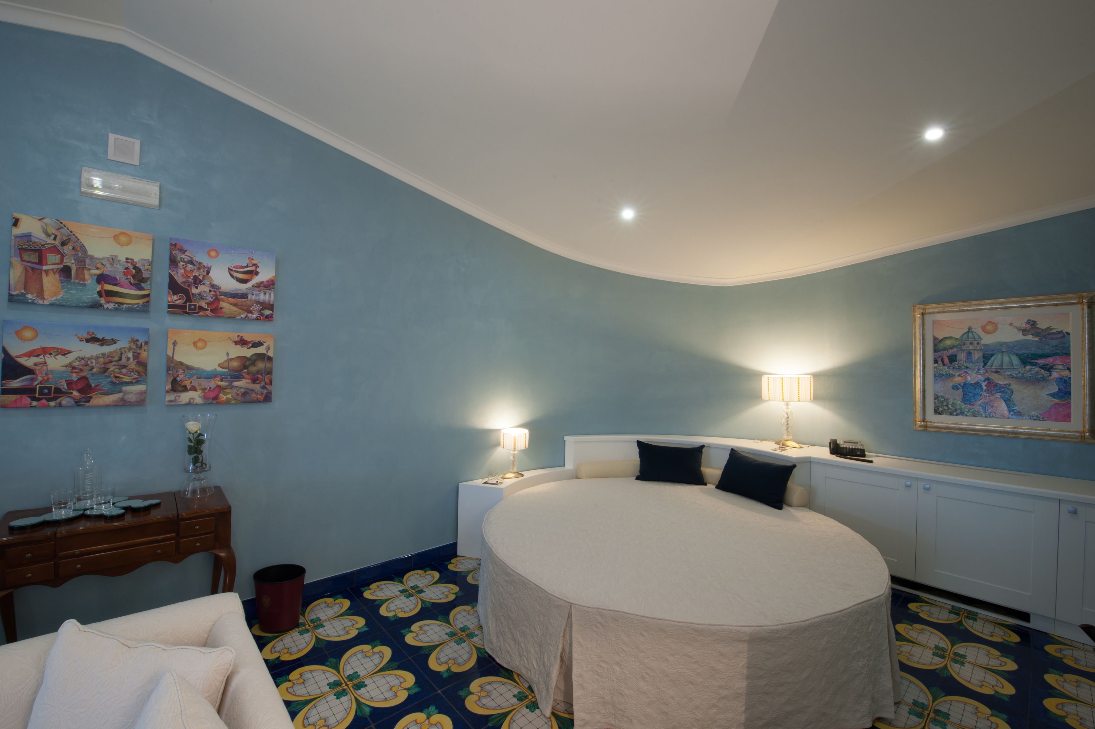A bedroom with a round bed, two bedside lamps, a nightstand, a dresser, and a wall with four framed pictures.