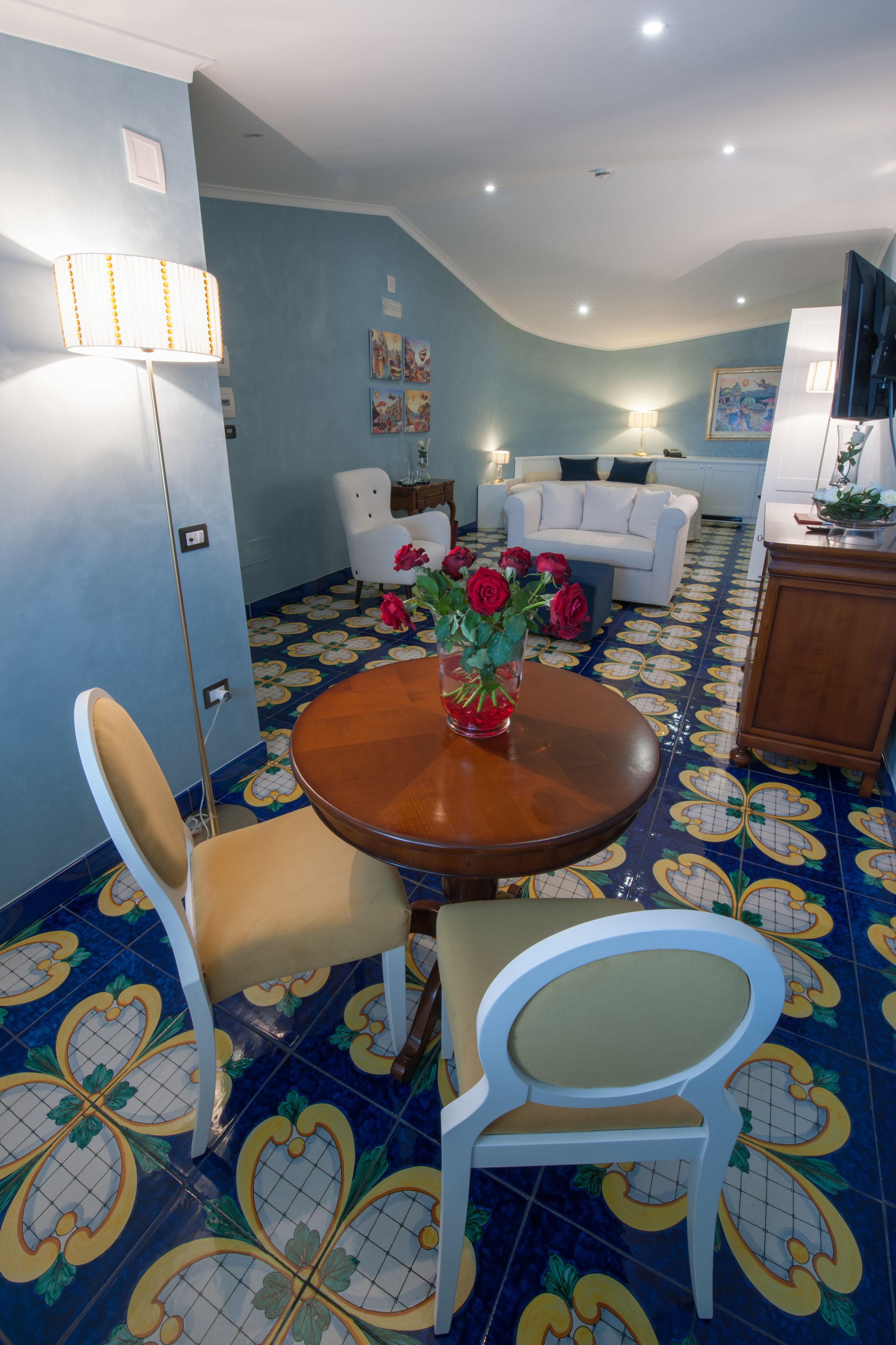 A dining area with a round wooden table, two white chairs, and a vase of red roses. The room has blue walls and a patterned floor.