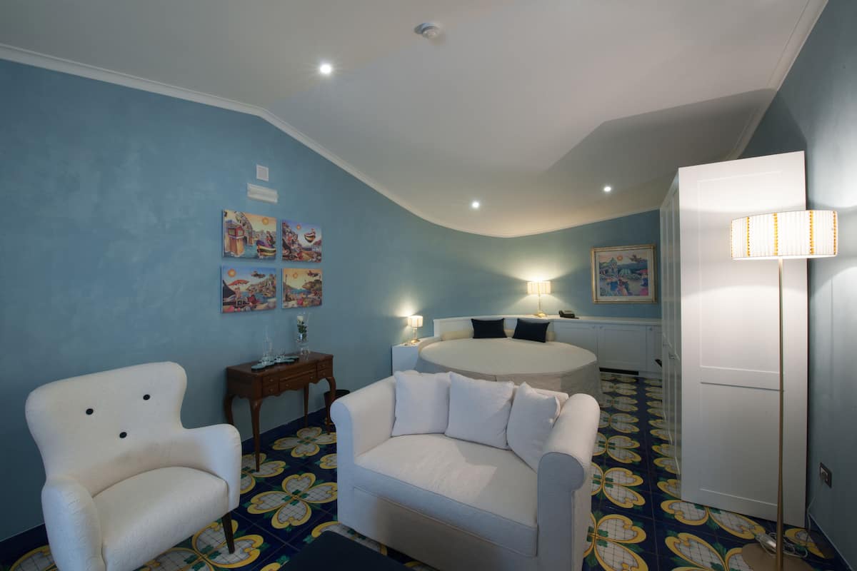 A room with a curved ceiling, a bed with a round headboard, a white armchair, a sofa, a small table with a lamp, and a door.