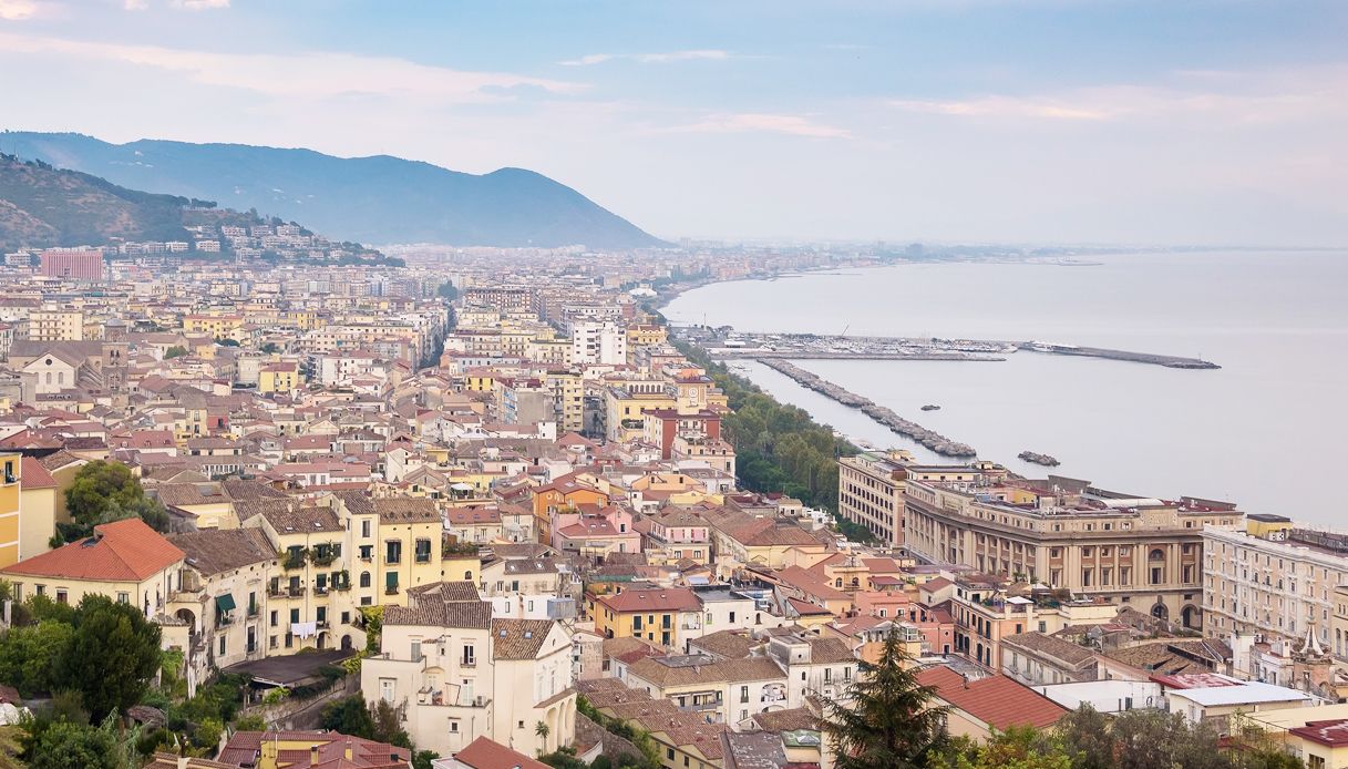 Un paisaje urbano costero con edificios densos, un paseo marítimo y una cadena montañosa distante.