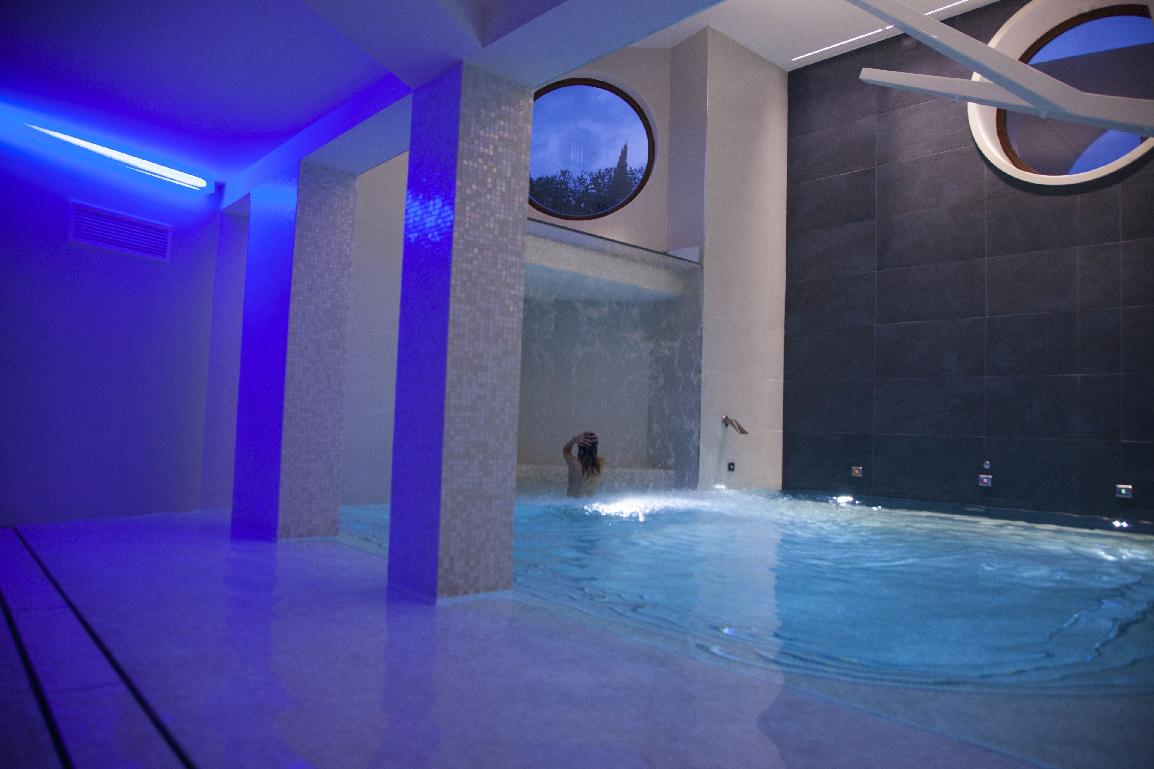 Une piscine intérieure moderne avec une vue sur une montagne, visible à travers une fenêtre circulaire, et entourée de murs en pierre et d’un éclairage bleu.