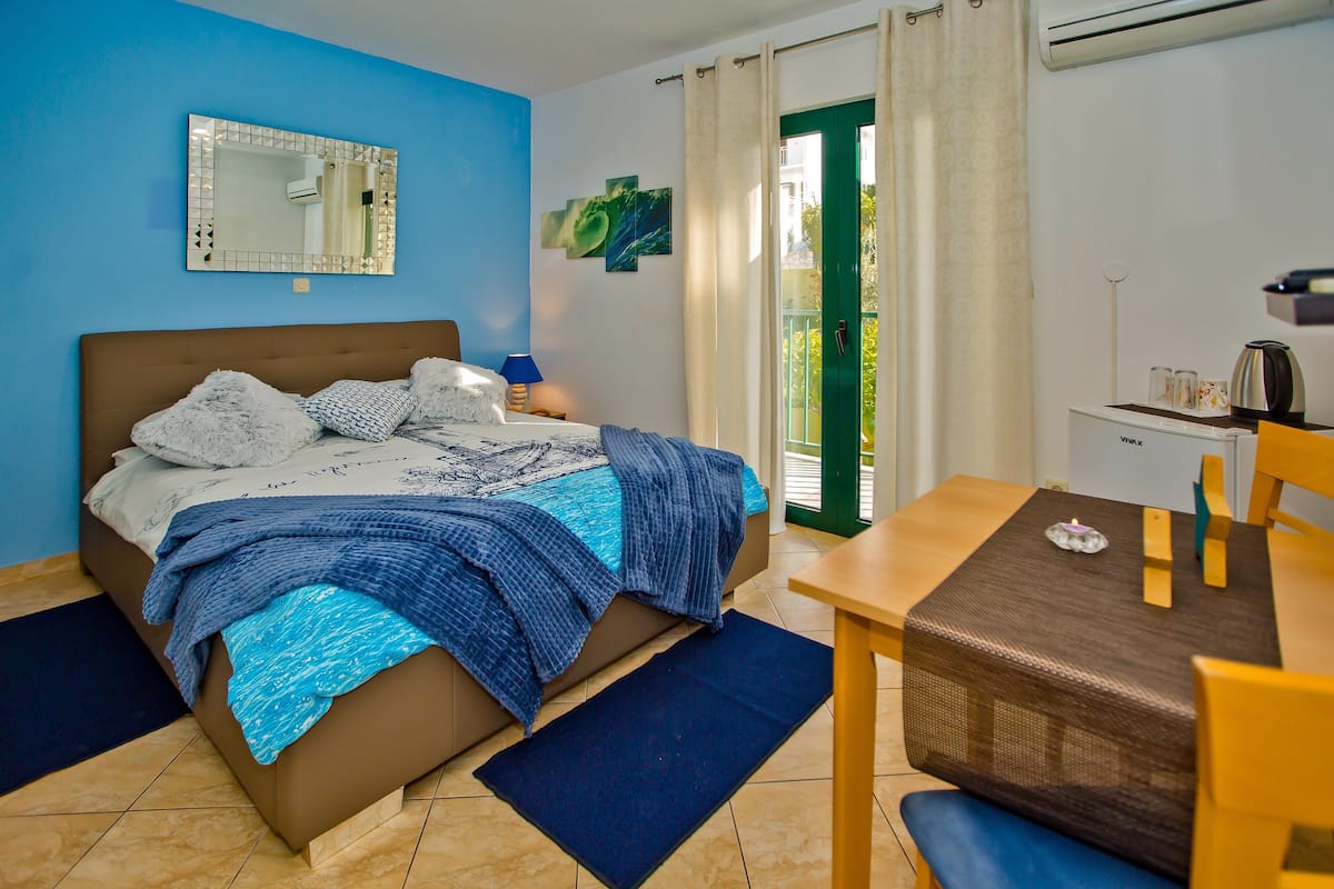 A bedroom with a bed, a desk, a chair, a mirror, and a view of the outdoors.