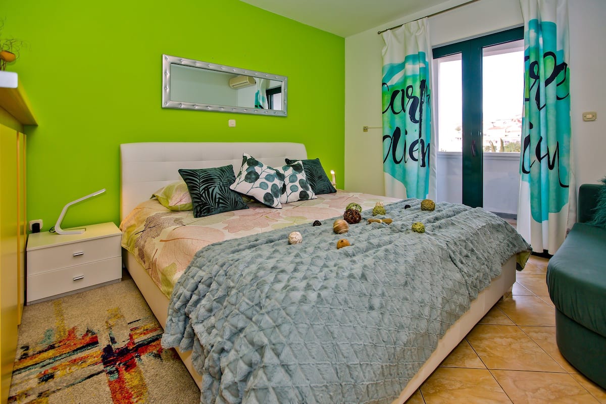 A bedroom with a bed, a nightstand, a mirror, a window with curtains, and a green wall.
