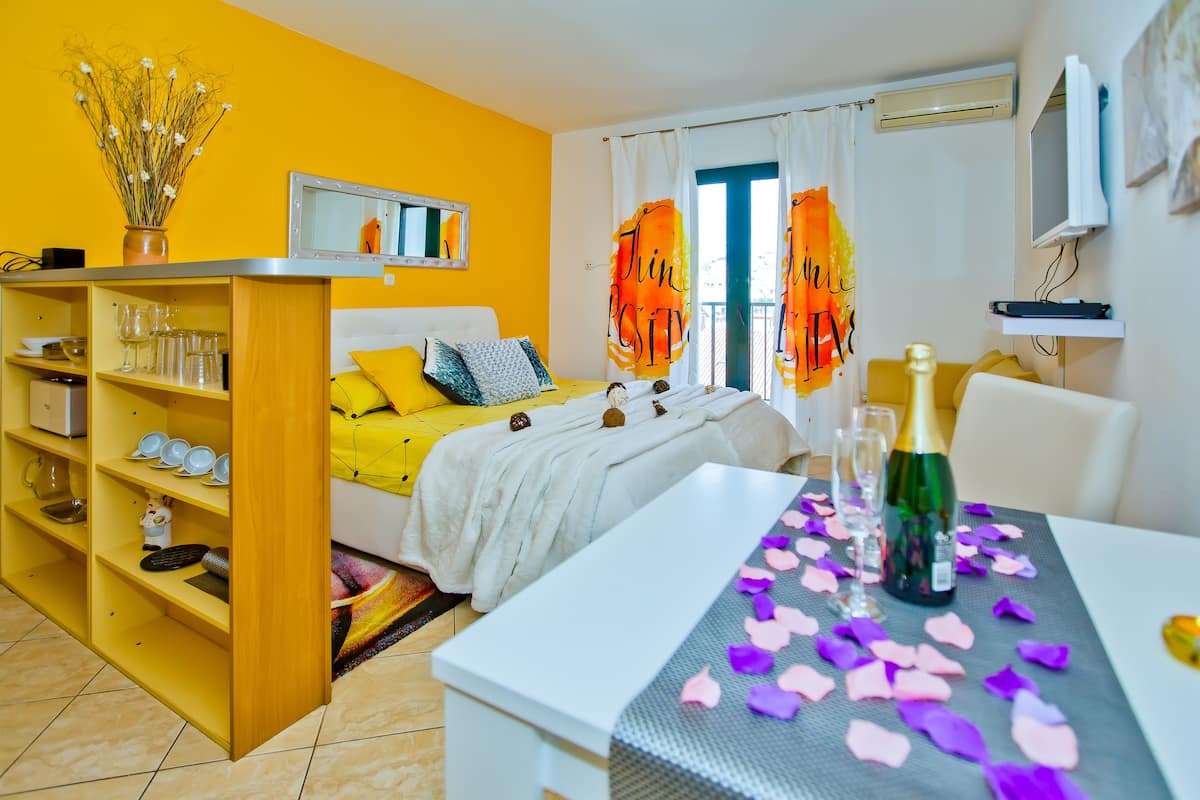 A bright yellow room with a bed, a small table with a bottle and glasses, and a shelf with dishes.