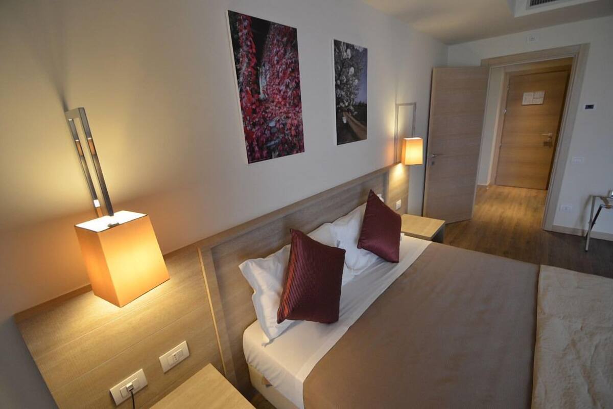 Una habitación de hotel con una cama, dos lámparas fijadas a la pared y dos cuadros enmarcados.