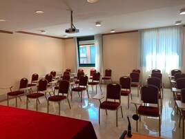 Un salón de conferencias con filas de sillas y mesas, un proyector y un amplio ventanal con cortinas.