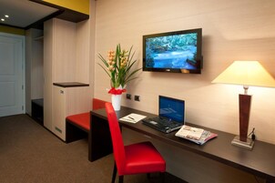 A hotel room with a desk, a laptop, a television, a red chair, a vase with flowers, and a lamp.