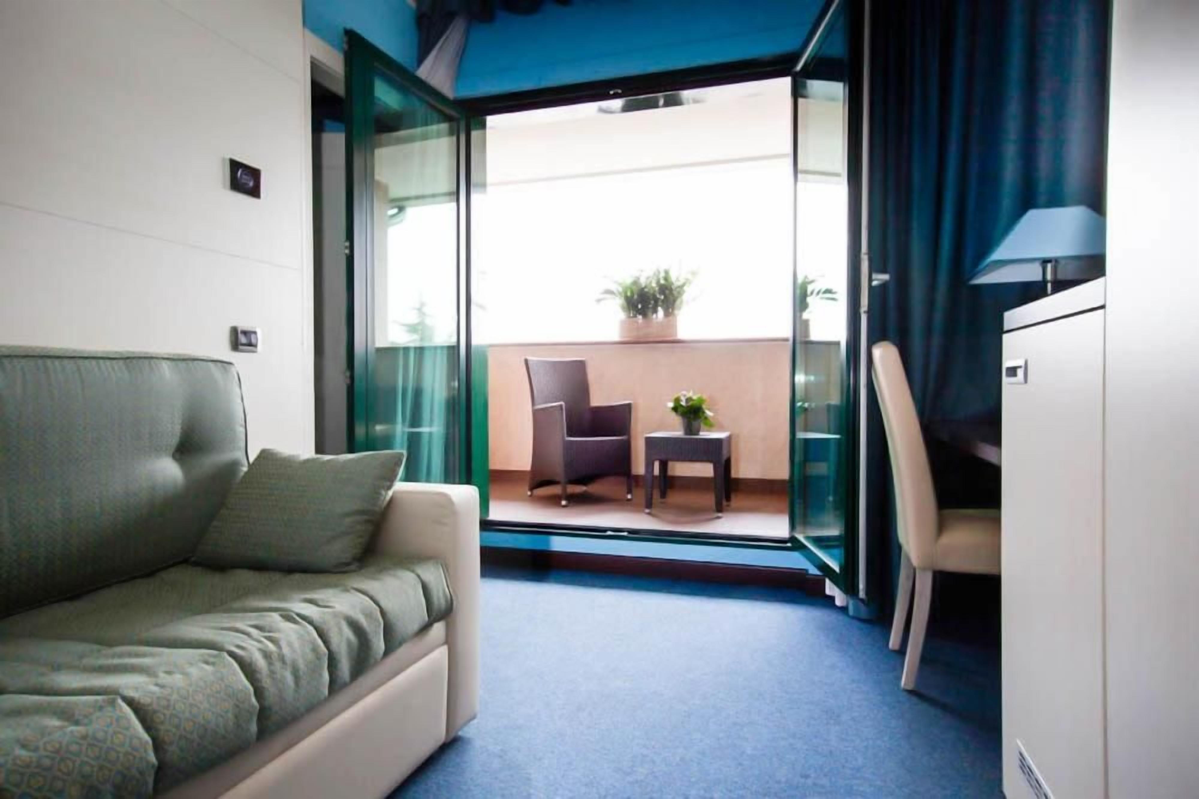 A hotel room with a sofa, a chair, a small table, and a balcony with a potted plant.