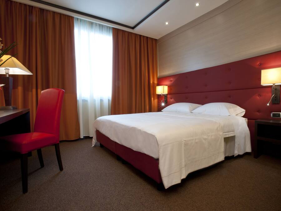 Habitación de hotel con una cama grande, un escritorio, una silla, dos lámparas de noche y un ventanal con cortinas.