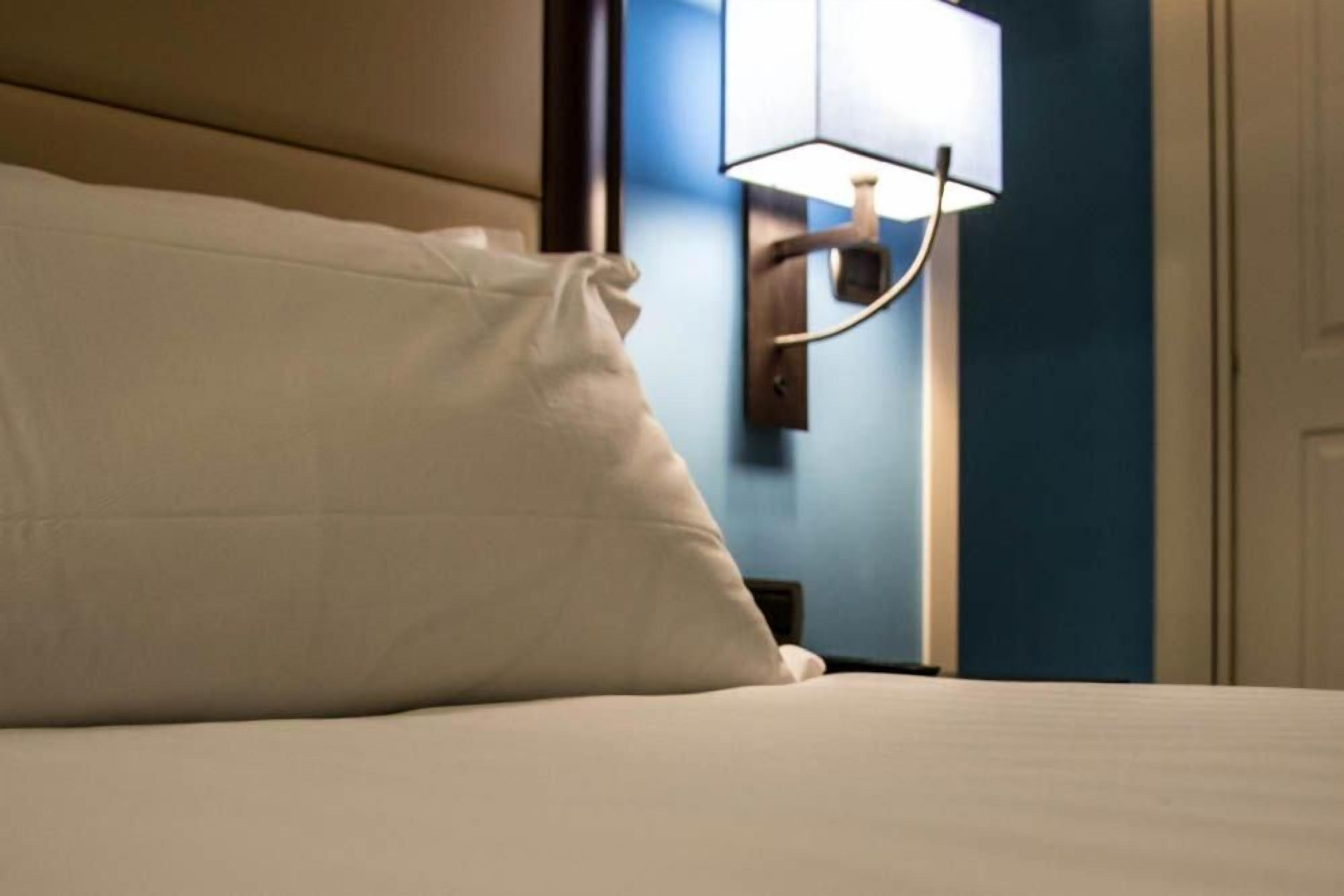 Una cama con almohada blanca y lámpara de pared.